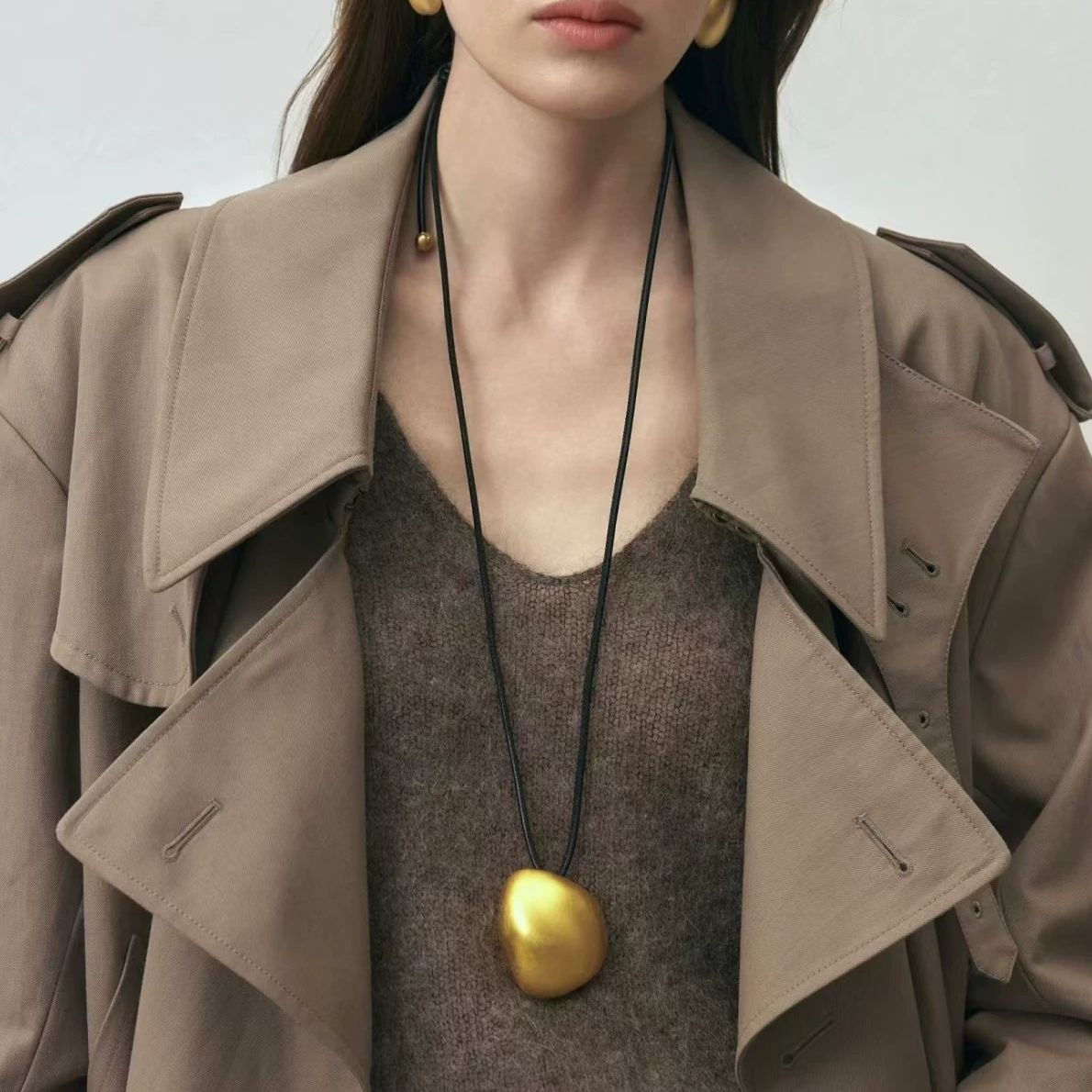 Model wearing a gold-tone sculptural pebble pendant necklace on a long dark cord with a taupe trench coat